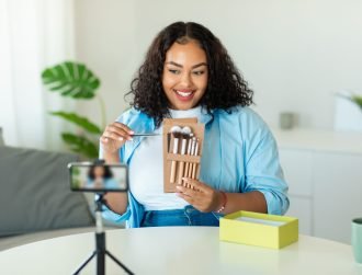 Beauty Blogging. Black Blogger Lady Making Makeup Video Tutorial For Her Blog Filming On Cellphone And Showing Cosmetic Brushes Sitting At Desk At Home. Selective Focus